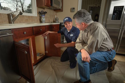 plumber in blue uniform explaining plumbing work under kitchen sink